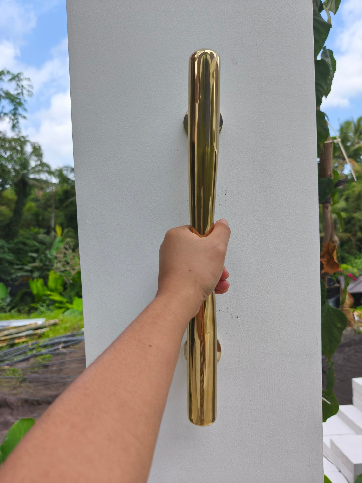 Hand gripping a solid brass door pull handle outdoors in a tropical garden, large gold entry door pull with a comfortable tapered grip for front doors
