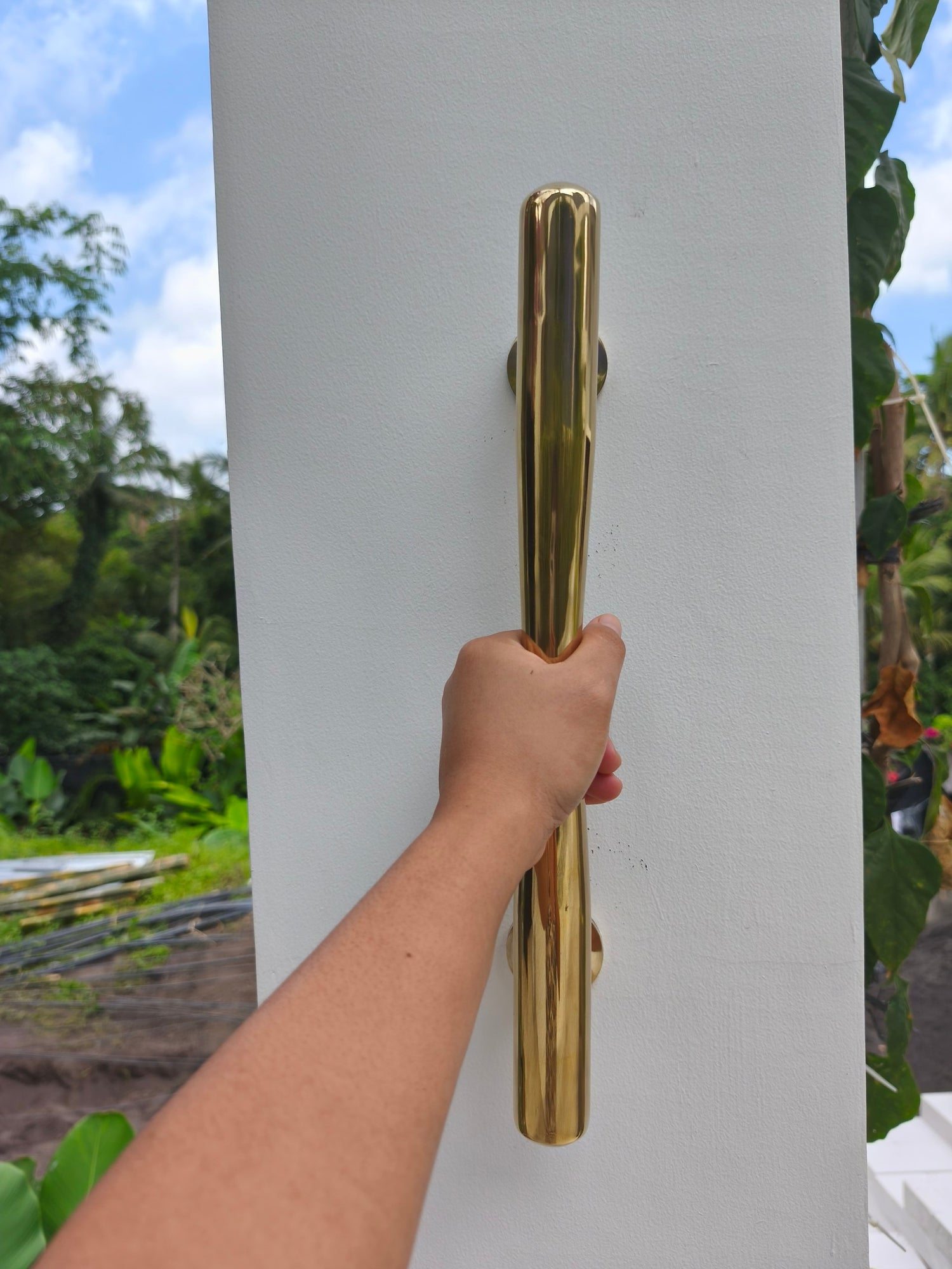 Polished brass door pull held upright in a garden setting, 40 cm gold door handle for Bali style villas, tropical homes and garden gate entrances