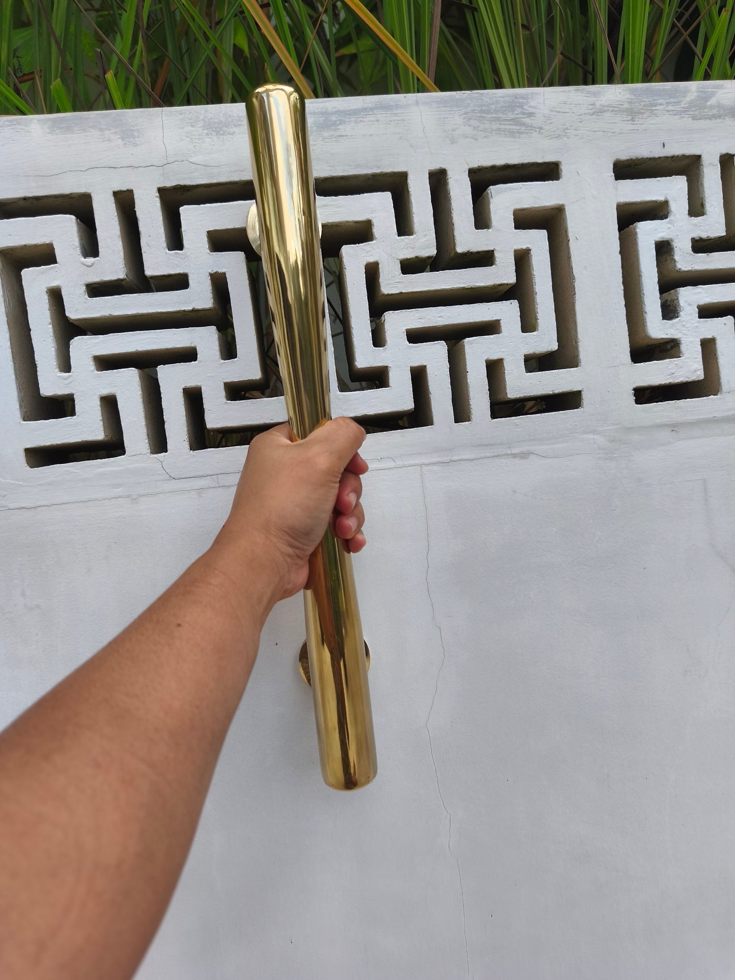 Hand holding a 40 cm polished brass door handle against a patterned white wall, large gold door pull for villas, hotels and modern entrances